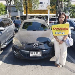 ภาพผลงานรับซื้อรถเก่า Mazda 2 โฉม DE ลูกค้ายืนถ่ายภาพคู่กับรถและป้ายโลโก้ CAR2GOLD