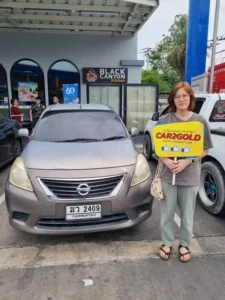 ลูกค้ายืนถือป้าย CAR2GOLD ถ่ายคู่กับรถเก่า Nissan Almera โฉม N17 หลังตกลงขายรถให้ทางร้าน